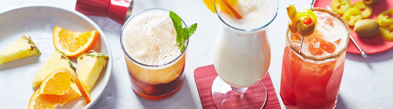 Three mocktails - a pineapple drink, non-alcoholic caesar and non-alcoholic moscow mule - on a white marble surface.