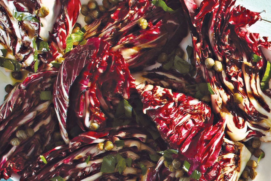 A white serving bowl on a blue background with grilled purple and white radicchio topped with a caper dressing.