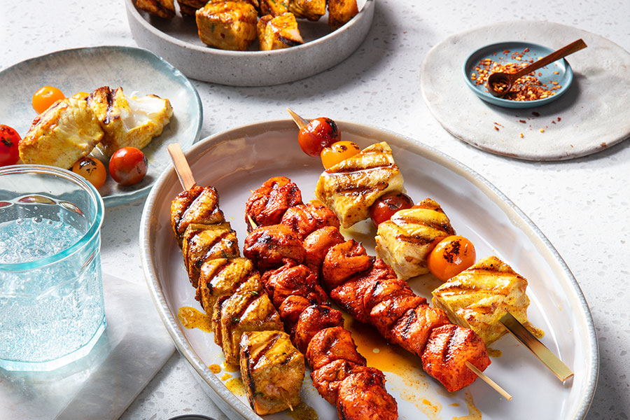 Three Indian-inspired kabobs including flaky white fish, chicken and blistered tomatoes with crushed chilis off to the side in a blue dish.