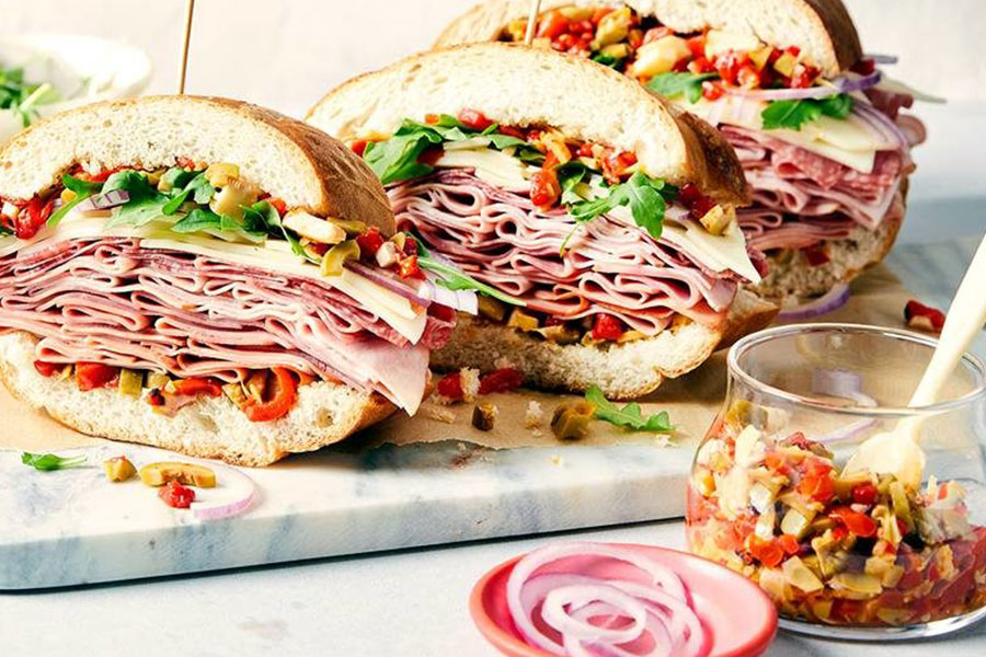 Deli meat, cheese, olive, roasted peppers, red onion, and arugula on a French loaf, sitting on a marble chopping block with next to a bowl of chopped, pickled vegetables.