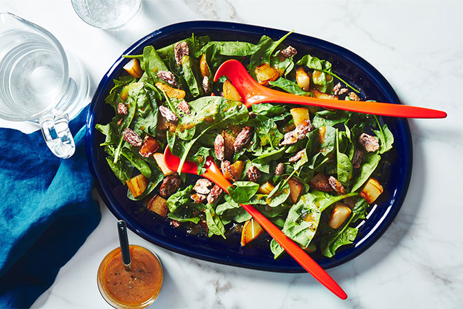 Blue platter with green spinach salad with chunks of roasted pears and pecans, and two red salad tongs.