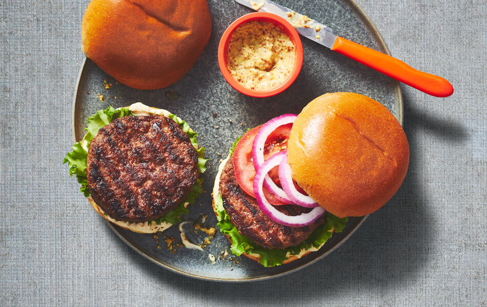 Overhead picture of gray plate with open Canadian cheddar chuck beef burger with toppings next to ramekin of mustard.