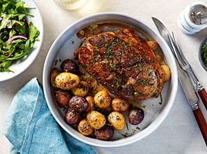 Boneless leg of lamb roasted in a white baking dish alongside roasted new potatoes on a white countertop, next to a fresh green salad.