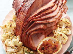 Glazed, sliced Panache hickory smoked honey spiral ham on a white platter with roasted cauliflower on the side, next to a carving fork and knife.