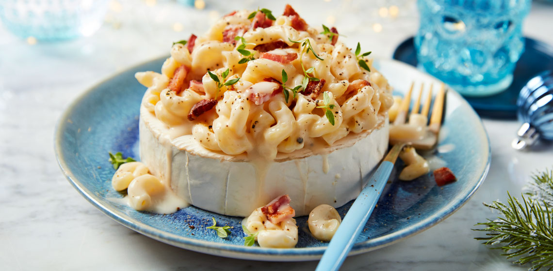 Light blue plate on white holiday tabletop with a wheel of brie mac nâ€™ cheese on top, with thyme garnish and a fork on the side.