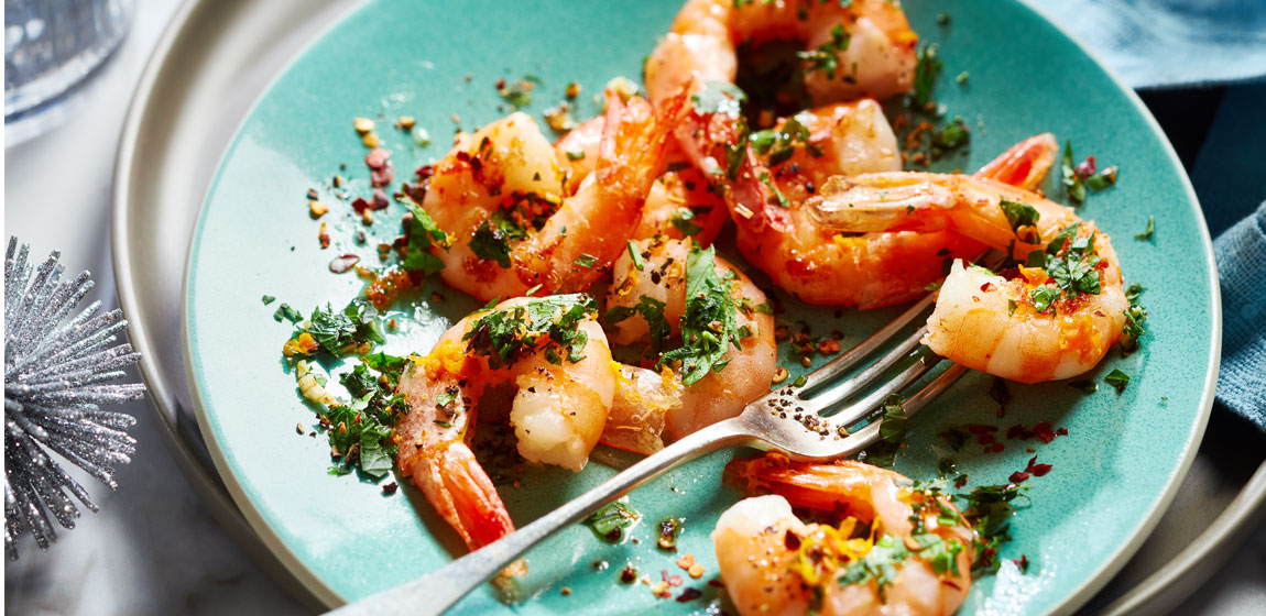 Blue plate with shrimps from Compliments Naturally Simple Cooked Pacific White Shrimp shrimp ring, dressed in green chili and orange gremolata on a white tabletop.