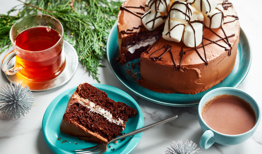 White marble surface with a whole hot chocolate cake with marshmallows on top and one slice cut out and a blue side plate with cake slice and fork.