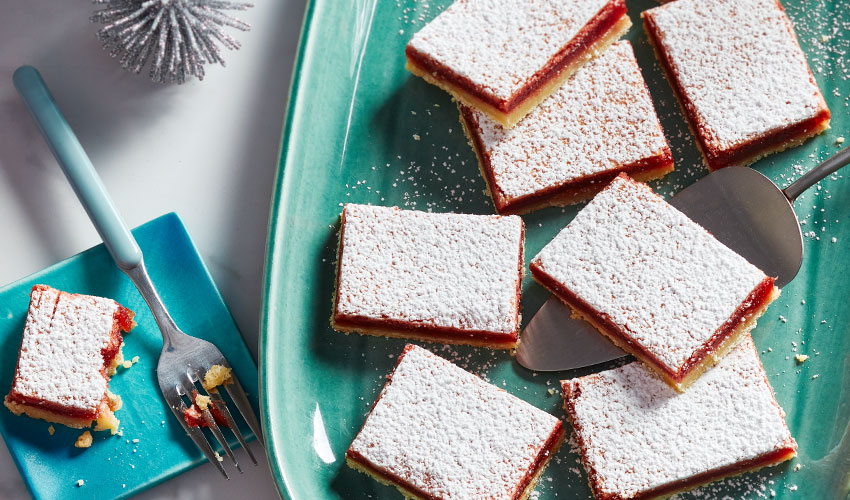 large green squared corned oval platter with lemon-cranberry squares, topped with icing sugar and a metal serving spatula.