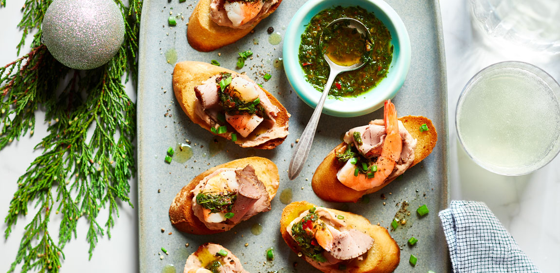 Large rectangular grey platter with shrimp and roast beef crostini, on a white marble tabletop with festive dÃ©cor and evergreen around the edge of the image.