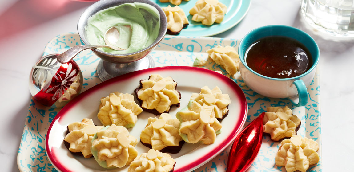 Marble tabletop with a blue and white tray topped with a plate of whipped shortbread cookies dipped in chocolate, and a short cup of tea, with some holiday ornaments for decoration.