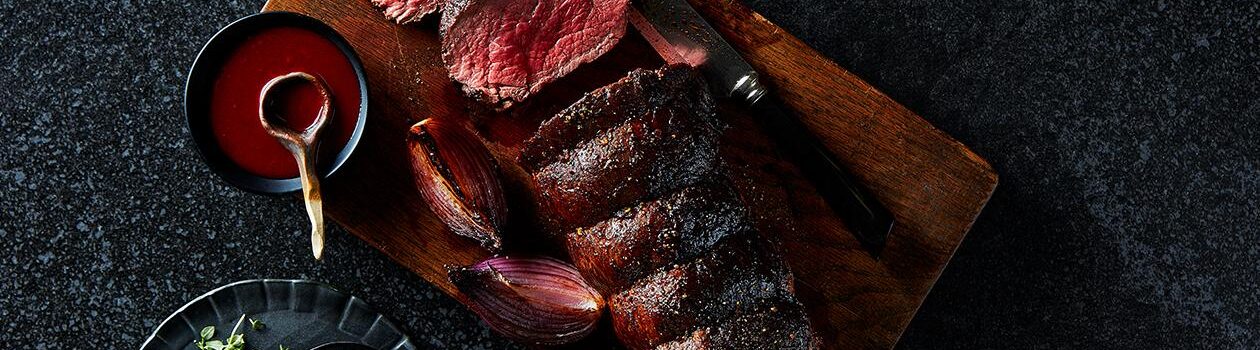 A whole roasted Sterling Silver beef tenderloin on a cutting board, with a few slices cut next to a small bowl of sauce with a wooden spoon.