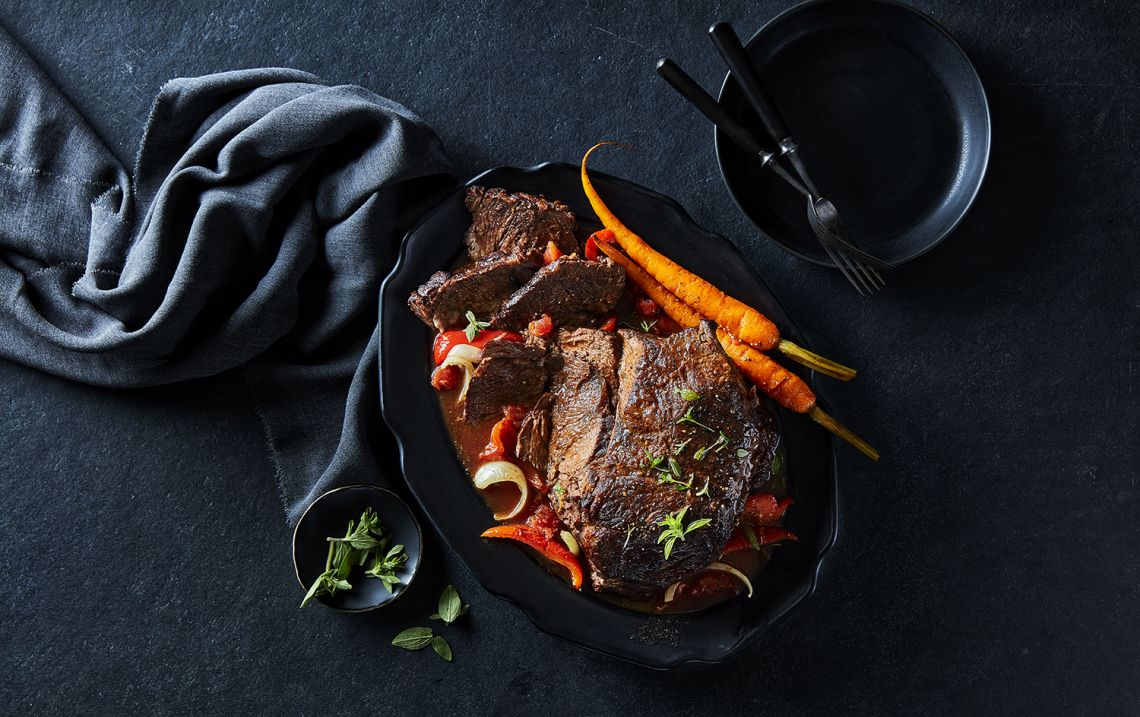 Dark charcoal table surface with a black oval serving platter of sirloin roast with roasted carrots and onions.