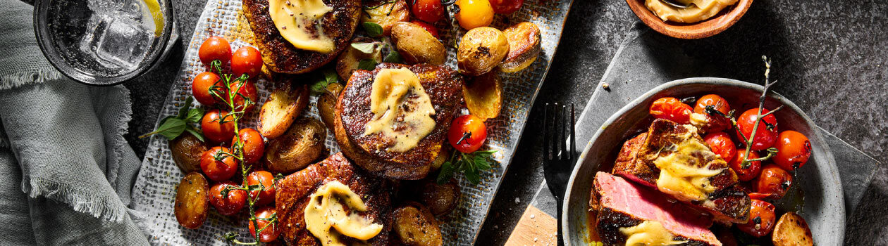 Two Shawarma-Marinated Beef Tenderloins topped withTahini Butter on a gray platter served with blistered tomatoes and roasted baby potates.