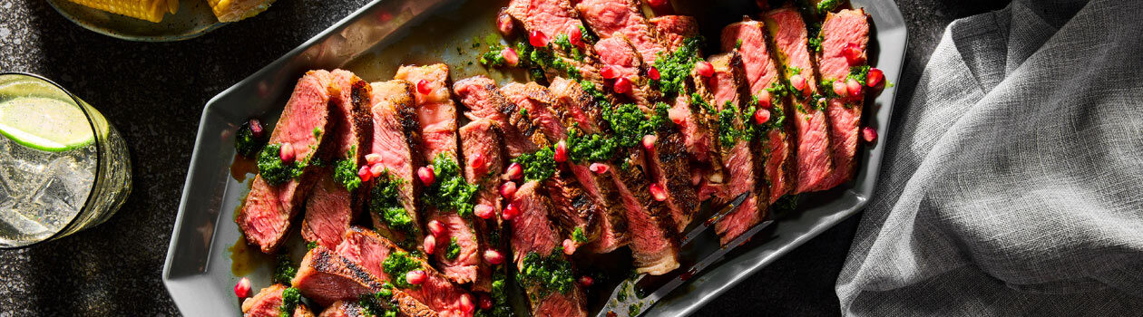 Grilled Syrian Seven Spice Rib Steak sliced on a grey platter with a nearby bowl full of green, herbaceous Chermoula and a bowl of grilled corn.