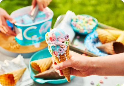 young hand holding waffle ice cream cone with unicorn twirl ice cream studded with fruity hoops cereal around the edges.