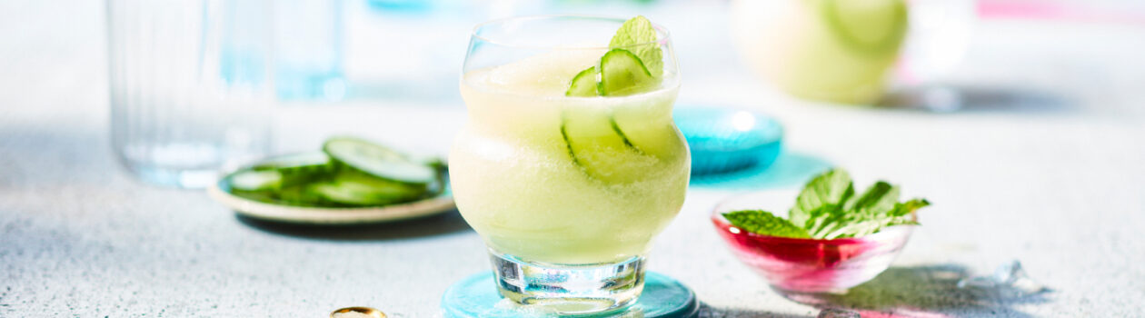 Low highball glasses of the Cucumber Gimlet Slush that’s a pale green sitting on top of a turquoise coaster on a gray countertop.