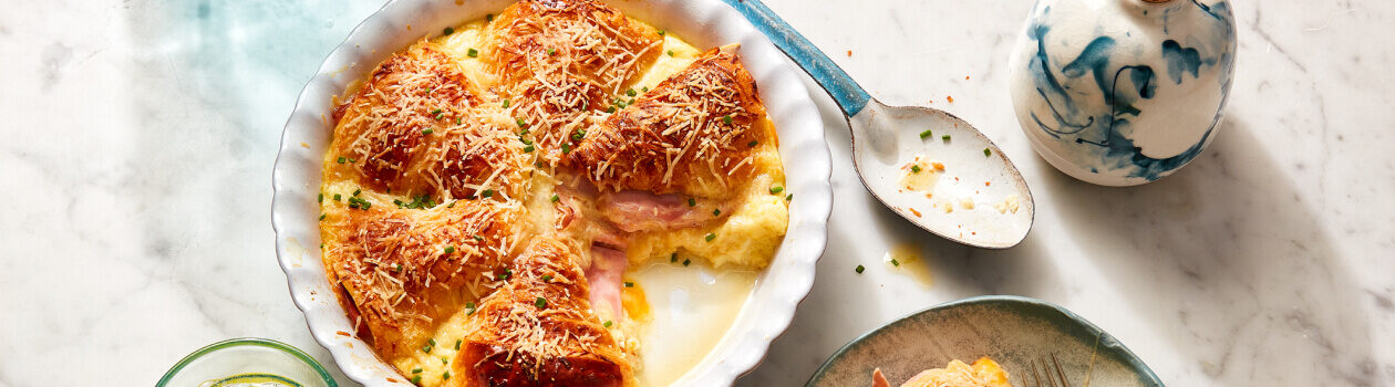 Ham and Cheese Croissant Strata in a white baking dish with a serving next to it on a gray dish with a side salad.