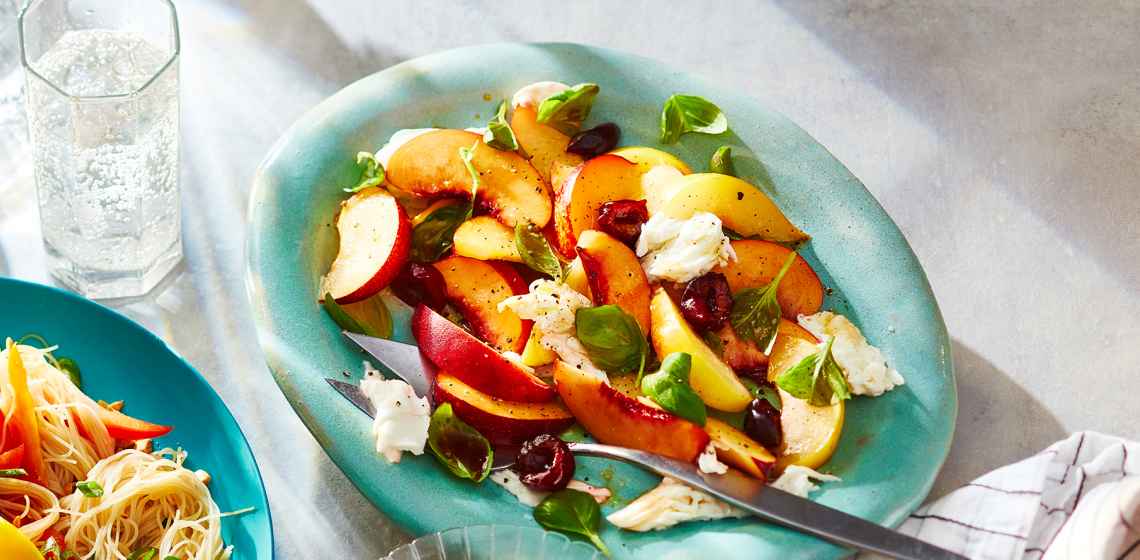 Light green serving platter topped with a fresh, stone fruit Caprese-style salad and serving spoons.