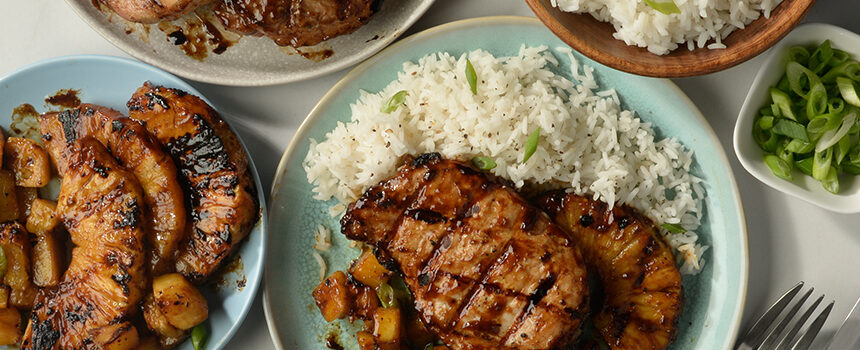 A blue plate is filled-out to the rim with fluffy white rice topped with grilled pineapple and a cross-hatch grilled pork chop.