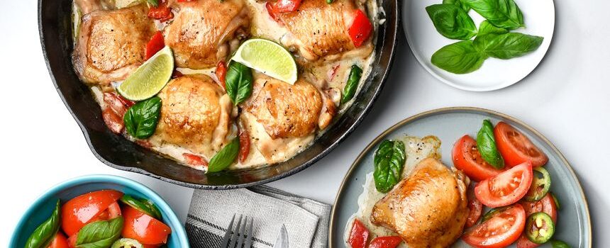 A non-stick pan filled with Thai green curry chicken thighs and tomato salad, served with slices of lime.