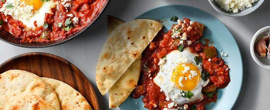 Baked Shakshuka eggs in a tomato sauce nestled into a cast iron pan, next to a plate of shakshuka with mini naan bread on the side.