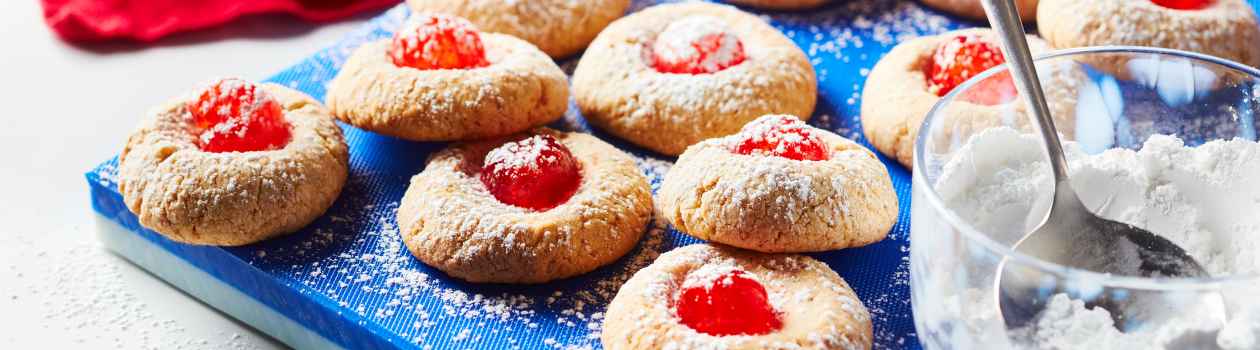 Blue cutting board with many cheesecake thumbprint cookies with red cherries on top and a dusting of icing sugar