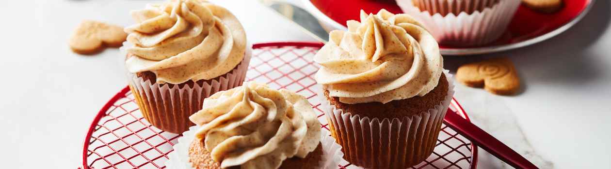 White marble surface with red cooling rack of three iced gingerbread cupcakes with brown butter buttercream, and a red plate in backgroud with extra cupcakes and a few scattered decorative ginger cookies