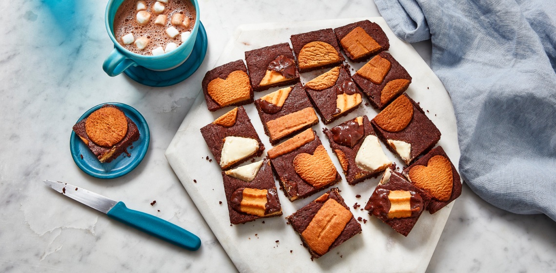 white marble surface with white cutting board of cut up brownies, studded across the top with Panache Belgian Biscuit cookie assortment