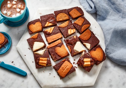 white marble surface with white cutting board of cut up brownies, studded across the top with Panache Belgian Biscuit cookie assortment