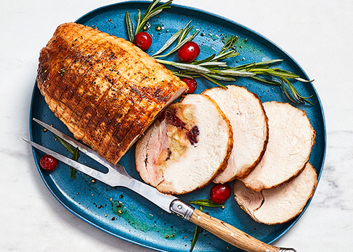 Stuffed turkey breast on blue serving dish with carving utensils, rosemary and cranberries.