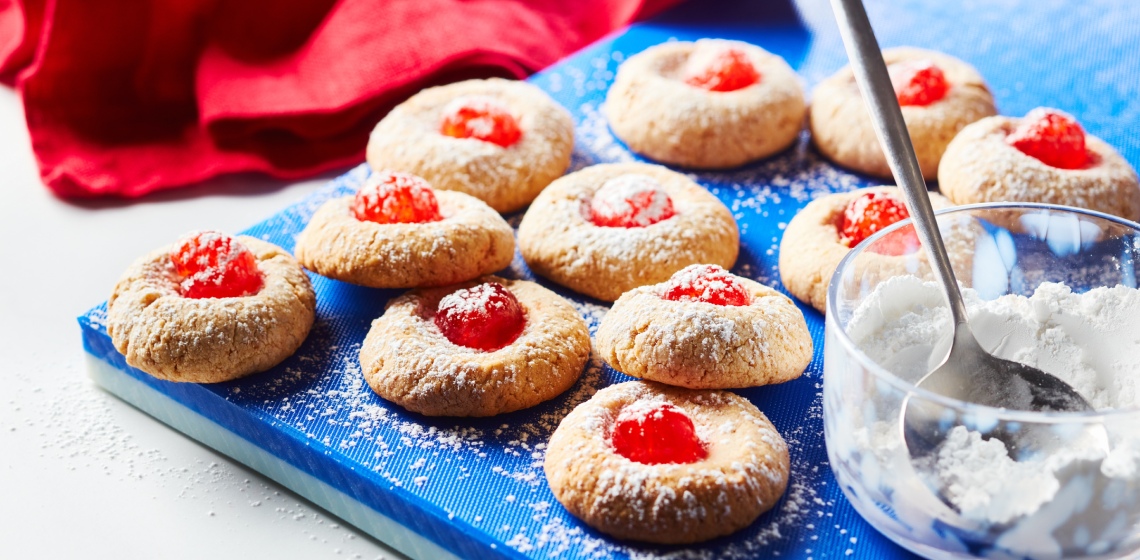 Blue cutting board with many cheesecake thumbprint cookies with red cherries on top and a dusting of icing sugar