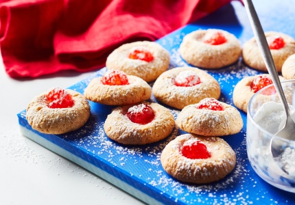 Blue cutting board with many cheesecake thumbprint cookies with red cherries on top and a dusting of icing sugar