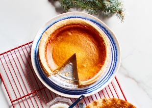 White marble surface with red cooling rack, holding one pumpkin pie with a slice cut out, and one strawberry rhubarb pie with a slice cut out