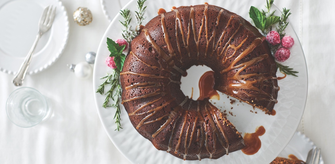 Overhead looking down at a white holiday tabletop with a white cake stand with a gingerbread Bundt cake drizzled with sauce, green and red garnish, and once slice cut out.