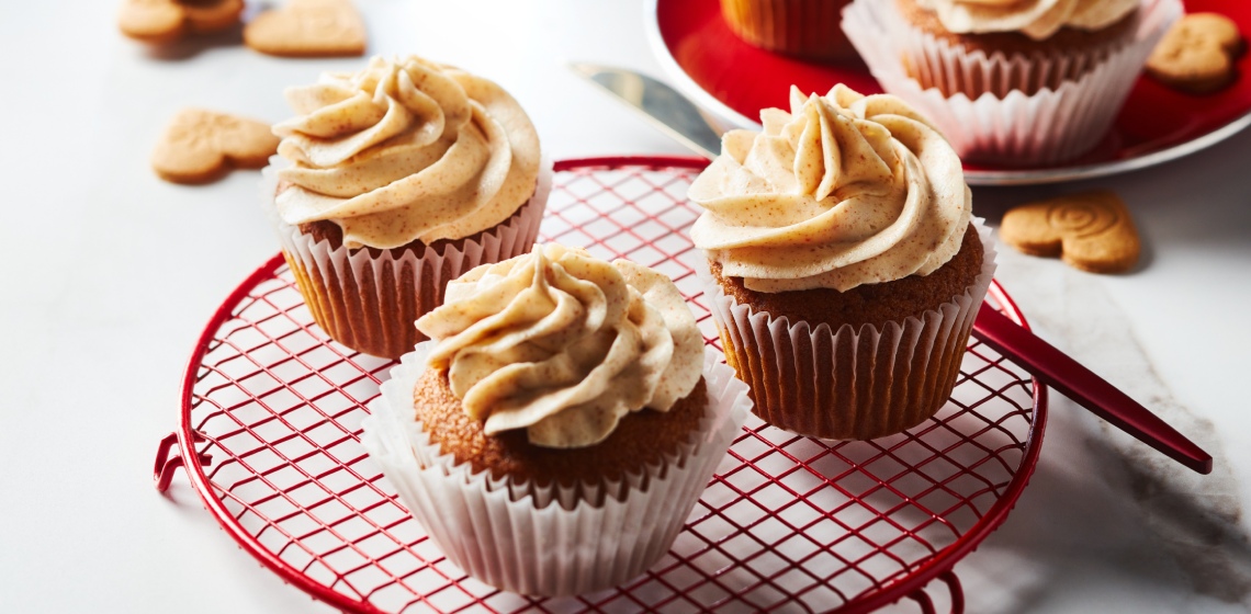 White marble surface with red cooling rack of three iced gingerbread cupcakes with brown butter buttercream, and a red plate in backgroud with extra cupcakes and a few scattered decorative ginger cookies