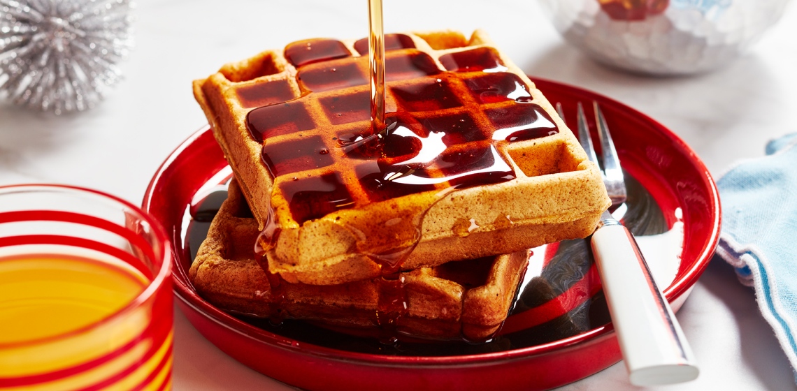 A close-up of waffles on a red plate with syrup being drizzled from up high, and a fork on the side.