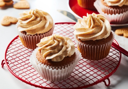 White marble surface with red cooling rack of three iced gingerbread cupcakes with brown butter buttercream, and a red plate in backgroud with extra cupcakes and a few scattered decorative ginger cookies