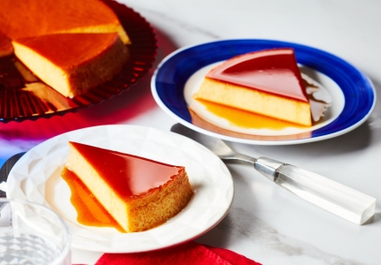 large red serving platter with caramel-topped round Mexican-style flan with two slices cut, one served out on blue-rimmed white side plate at right