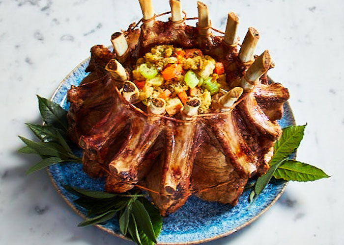 Crown roast on blue plate with green herb sprigs arranges around the edges.