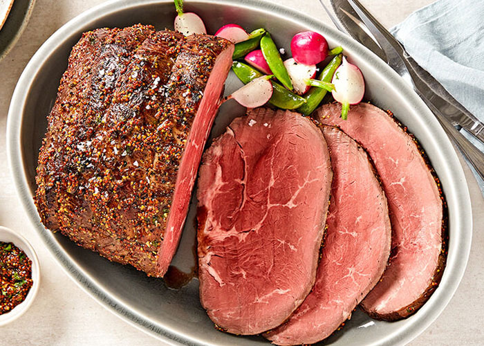 Top down view of roast sirloin and slices of roast sirloin on dish with snap peas and radishes placed beside. Bowl of sweet mashed potatoes is on the right and ramekin of spice blend on left