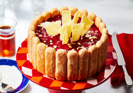 Red, orange and white cake stand with cranberry charlotte cake on top, with side plates ready to be served, and full tea cups to the side.