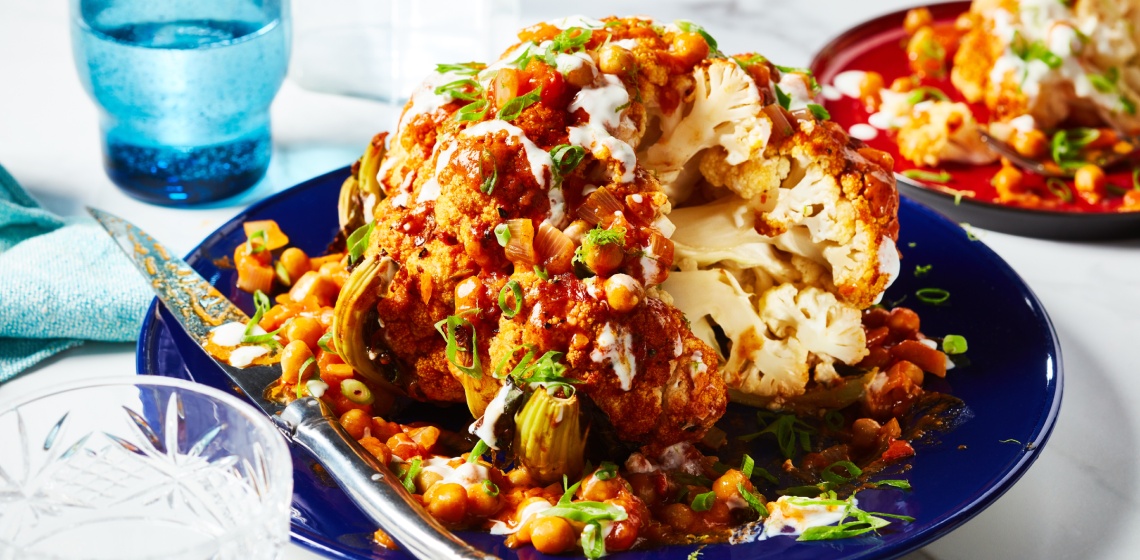 Round blue serving platter of whole-roasted tikka cauliflower with sauce overtop and slice cut out