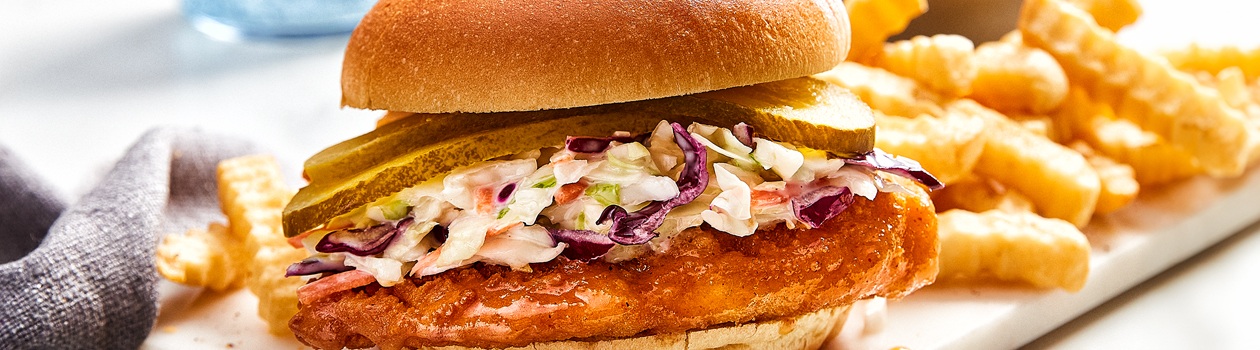 plate with a soft brioche bun layered with crispy fish, slaw and pickle slices, and a side of crinkle-cut French fries.