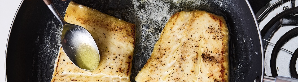 Black non-stick frying pan with two fish filets being pan seared in butter