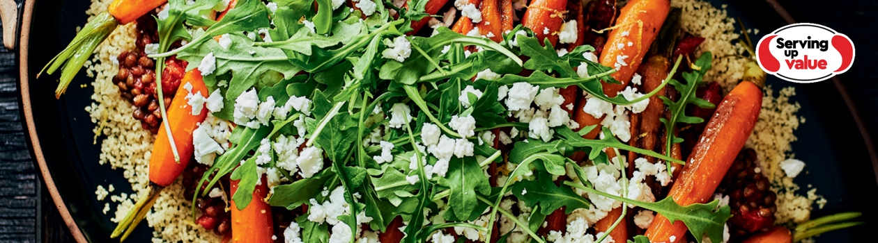 Large oval handled serving dish with couscous topped with lentils, roasted carrots, feta and fresh arugula.